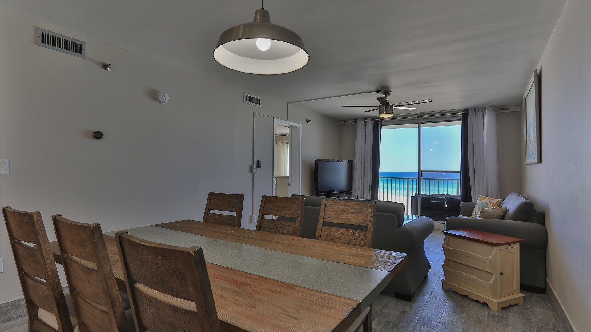 Un coin repas avec une table et des chaises en bois, un ventilateur de plafond, un téléviseur et une vue sur l’océan grâce à de grandes fenêtres.