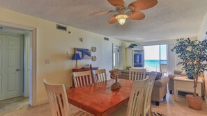 A dining area with a wooden table and chairs, a ceiling fan, and a view of the ocean through large windows. - Destin on the Gulf 606 (Destin)