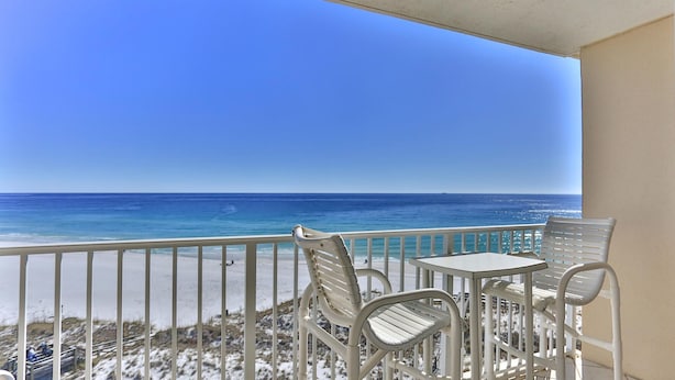 A balcony with white metal railings, two white metal chairs, and a small white table. The balcony overlooks a beach with clear blue water and a distant horizon. - Destin on the Gulf 706- Stunning Gulf Views-Pool (Destin)
