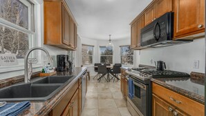 Cocina con armarios de madera, un lavamanos de acero inoxidable, estufa y microondas. Hay una mesa de comedor con sillas y una ventana con vistas a árboles.