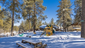 Un parque cubierto de nieve con un banco de madera y una mesa de picnic, rodeado de altos pinos.