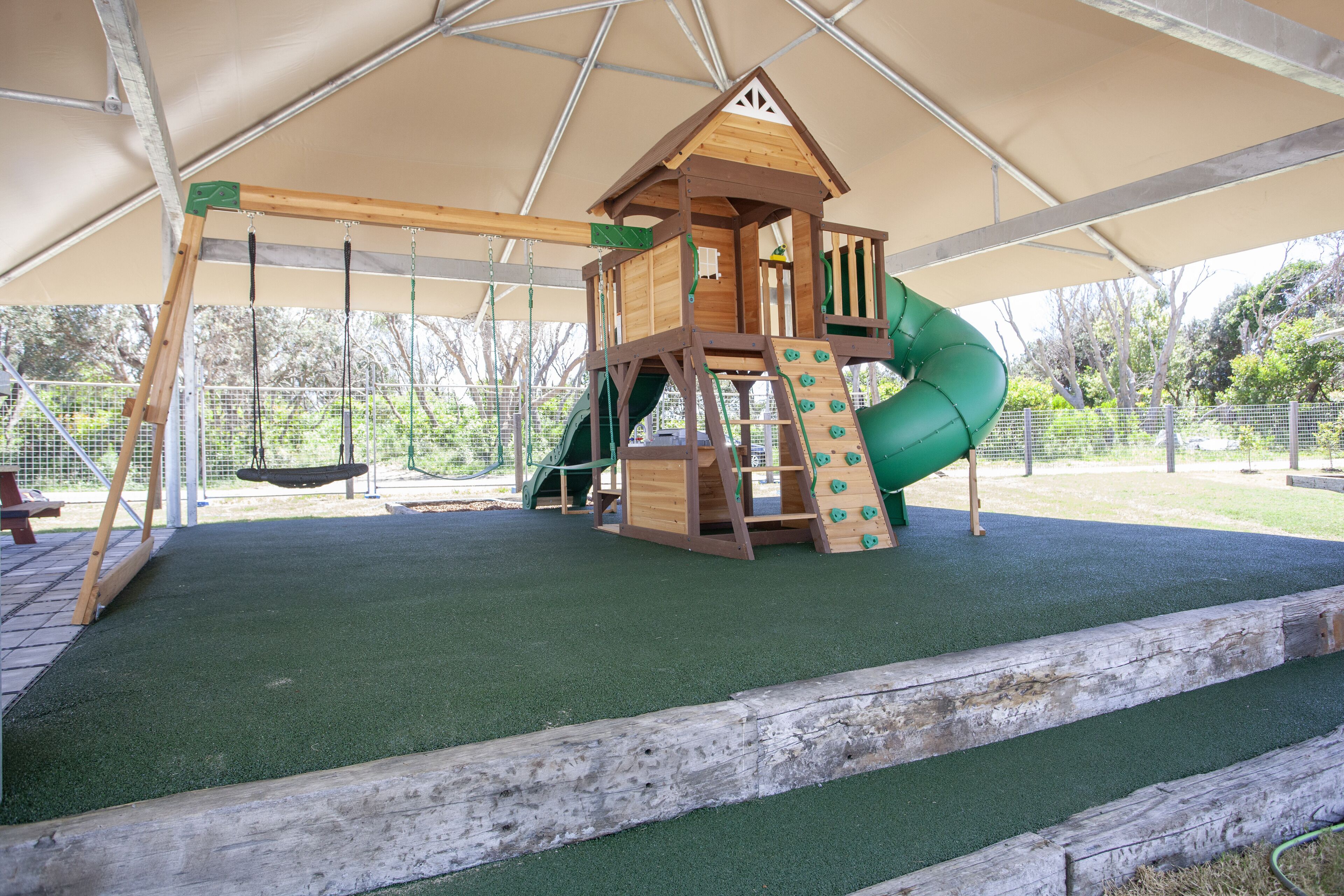 Estructura de juego de madera con tobogán y pared de escalada, rodeada por un área cubierta con césped artificial.
