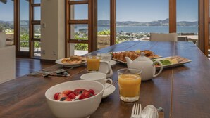 A breakfast table with croissants, fruit, and juice, overlooking a scenic view of the sea and hills.