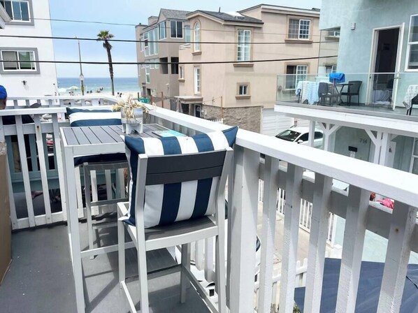 Un balcón con barandilla blanca, una mesa con sillas y vista a la playa y al océano.