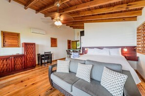 A cozy room with a wooden ceiling, a bed with white linens, a grey sofa with patterned cushions, a wooden cabinet, and a small table with chairs.