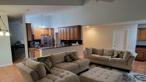 A living room with two sofas and a coffee table, a kitchen with wooden cabinets and a refrigerator, and a ceiling fan. - KING BED SUITE w. Large Backyard, 2 LVRMs & 2 FLs (Lauderhill)