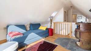 Een loft-achtige kamer met een blauwe bank, een rode poef, een houten vloer, een open haard en een trap naar een hoger niveau.