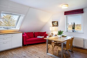 A room with a red sofa, a wooden dining table, a white cabinet, and a window with a view of trees. - Apartment "Voßberg Ferienwohnung 2" with Shared Garden & Wi-Fi (Grömitz)