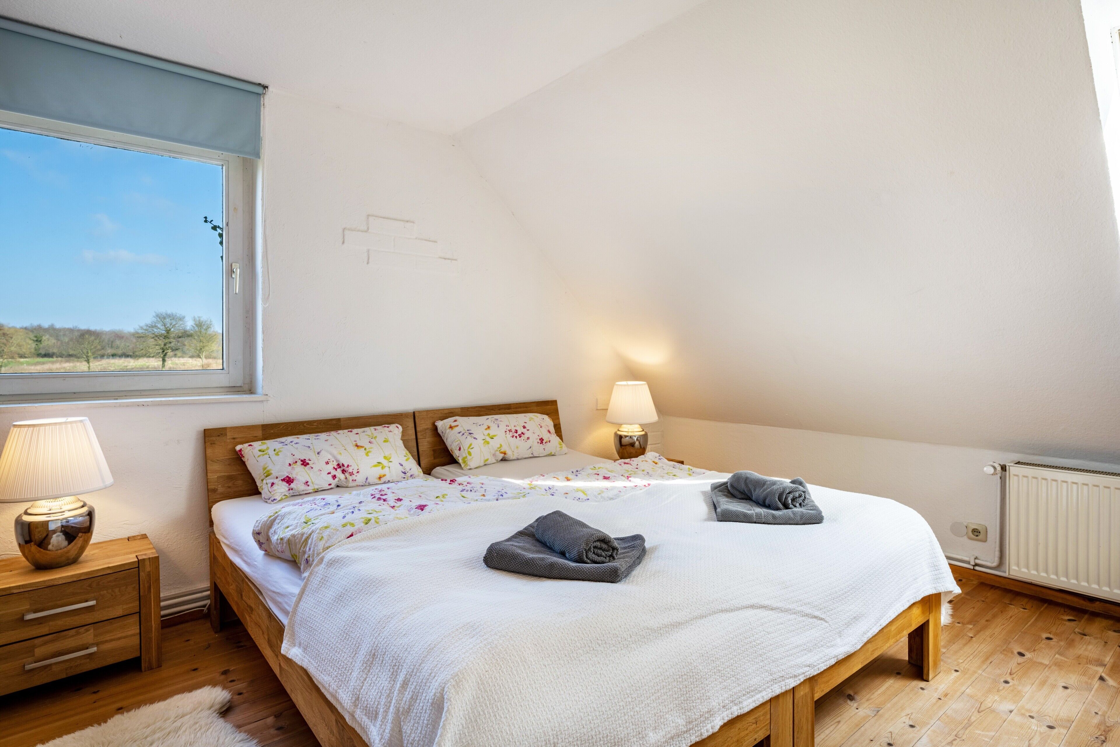 A bedroom with two beds, a window, a nightstand, a lamp, and a radiator.