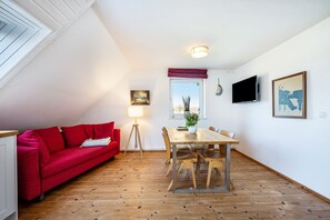 A room with a red sofa, a wooden dining table with chairs, a lamp, a TV, and a window with a view. - Apartment "Voßberg Ferienwohnung 2" with Shared Garden & Wi-Fi (Grömitz)