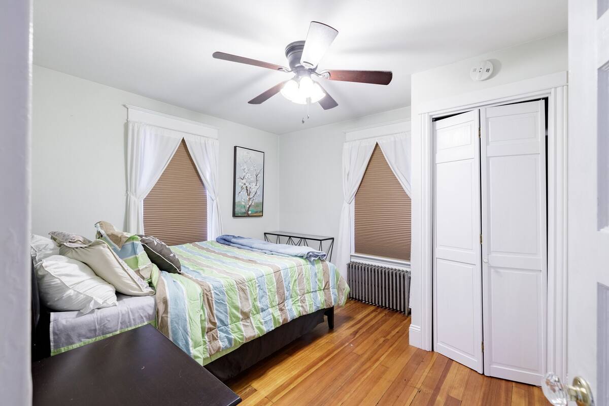 Une chambre à coucher avec un lit, un ventilateur de plafond, un radiateur et une armoire blanche.