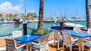 Un port de plaisance avec des bateaux, des palmiers et un restaurant aux tables bleues et aux chaises blanches.