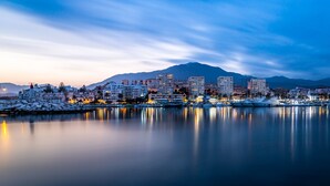 Un paysage urbain côtier au crépuscule, avec des bâtiments se reflétant sur des eaux calmes, une montagne en arrière-plan et une zone enneigée au premier plan.