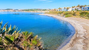 Un hôtel situé sur la côte, avec une mer d’un bleu limpide, une plage de sable fin et une vue sur la ville en arrière-plan.