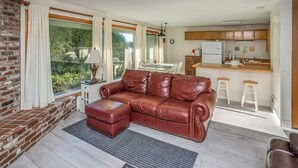 A living room with a leather sofa, ottoman, and a dining area with a table and chairs. A kitchen is visible in the background.