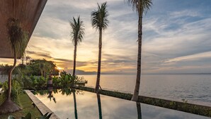 Une piscine à débordement paisible, offrant une vue dégagée sur le coucher de soleil sur l’océan, entourée de palmiers et d’une végétation luxuriante.