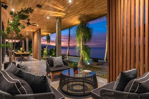 A modern indoor space with a dining area, seating, and a view of the ocean at sunset. - Sea View Beach Estate  (Buleleng)