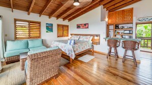 A cozy room with a bed, a sofa, a dining area with chairs, wooden ceiling beams, and a view of greenery outside.