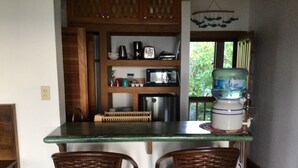 A kitchen with a green countertop, two wicker chairs, wooden cabinets, a water dispenser, and a window with a view of greenery.