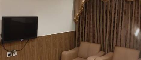 Une pièce dotée d’un téléviseur à écran plat fixé sur un mur en bois, de deux fauteuils beiges, d’un luminaire au plafond et d’une grande fenêtre avec des rideaux.
