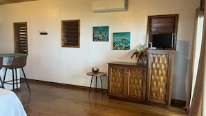 Una habitación con piso de madera, una mesa de comedor con sillas, una mesita redonda con un tazón, un aparador con puertas de mimbre y dos unidades de aire acondicionado fijadas a la pared.