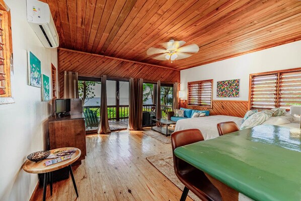 A spacious room with a wooden ceiling, a bed, a desk, and a balcony with a view of palm trees.