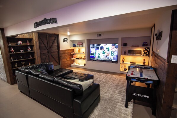 A living room with a black leather sofa, a foosball table, a TV, and shelves with trophies and memorabilia.