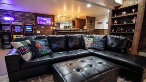 Un salon moderne avec un canapé d’angle, des consoles de jeux et un coin bar.
