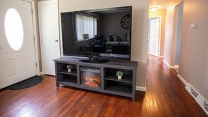 Un salon doté d’un grand téléviseur à écran plat fixé sur un meuble de télévision en bois foncé, d’une cheminée et d’un couloir menant à une autre pièce.