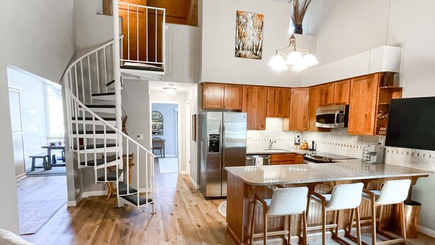Amplia sala de estar con escalera de madera, cocina equipada con electrodomésticos modernos y comedor con taburetes de bar.
