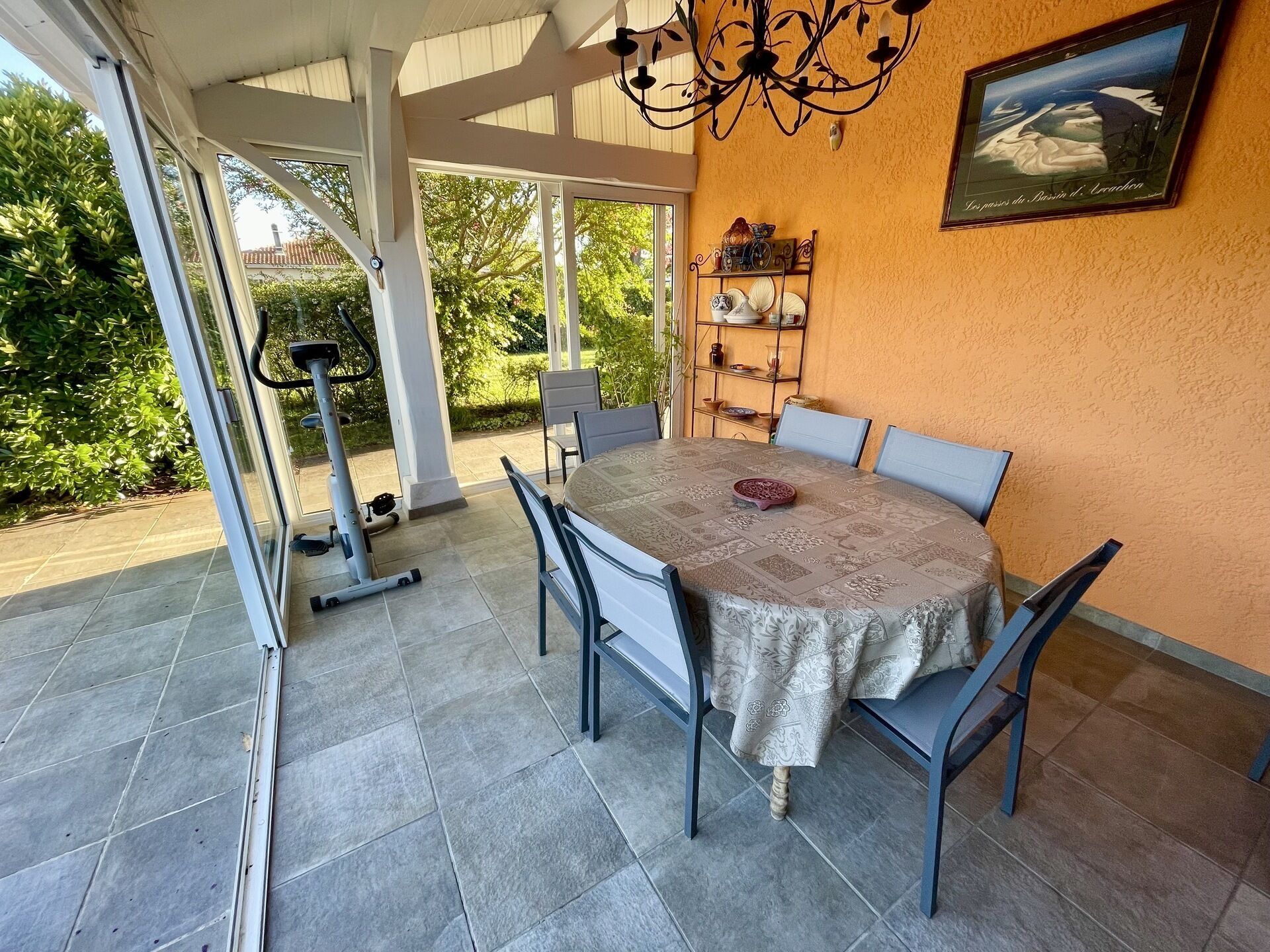 Un comedor con una mesa cubierta por un mantel estampado, rodeada de sillas. Se observa una caminadora al fondo, y una estantería con objetos decorativos en la pared.
