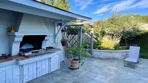 En uteplats med en eldstad i sten, en pergola med planterade växter och en sittgrupp.