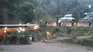 A rural setting with several small, traditional-style buildings with sloped roofs, surrounded by greenery and a dirt path leading to a covered area with lights. - Hidden Valley Camp (Nainital)