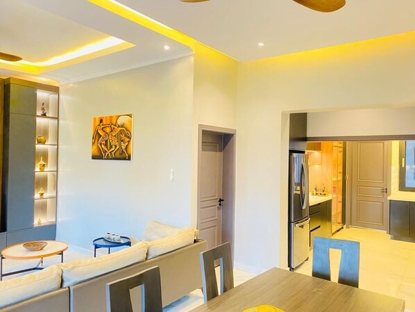 A modern living room with a sofa, dining table, and chairs. There is a kitchen area visible through an open door.