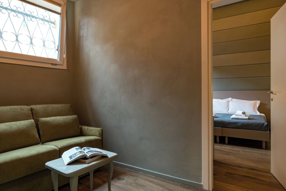 A room with a sofa, a small table with a book, and a bed with pillows and a blanket.