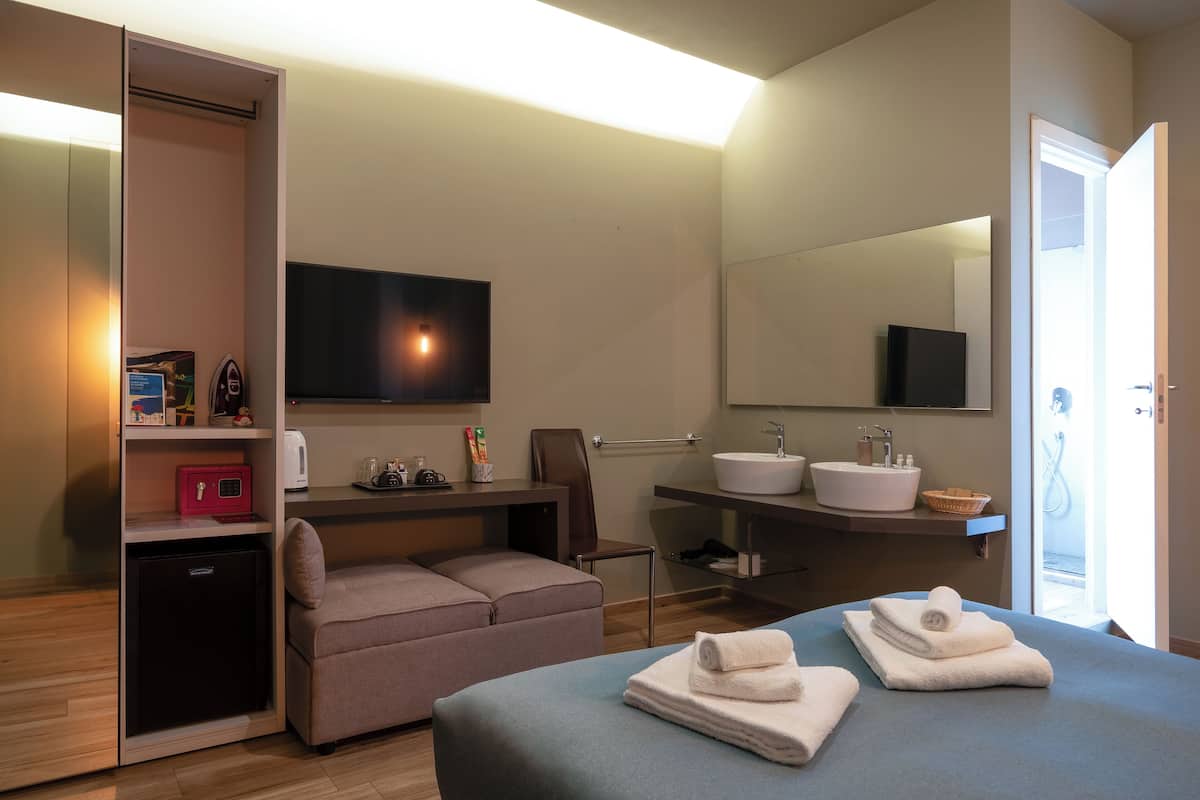 A modern hotel room with a flat-screen TV, a sofa, a small desk, a mini-fridge, a built-in sink, and a large mirror.