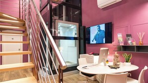 A modern interior with a staircase, a round table with chairs, a TV on the wall, and a glass elevator. - Gogò Petit Hotel (Florence)