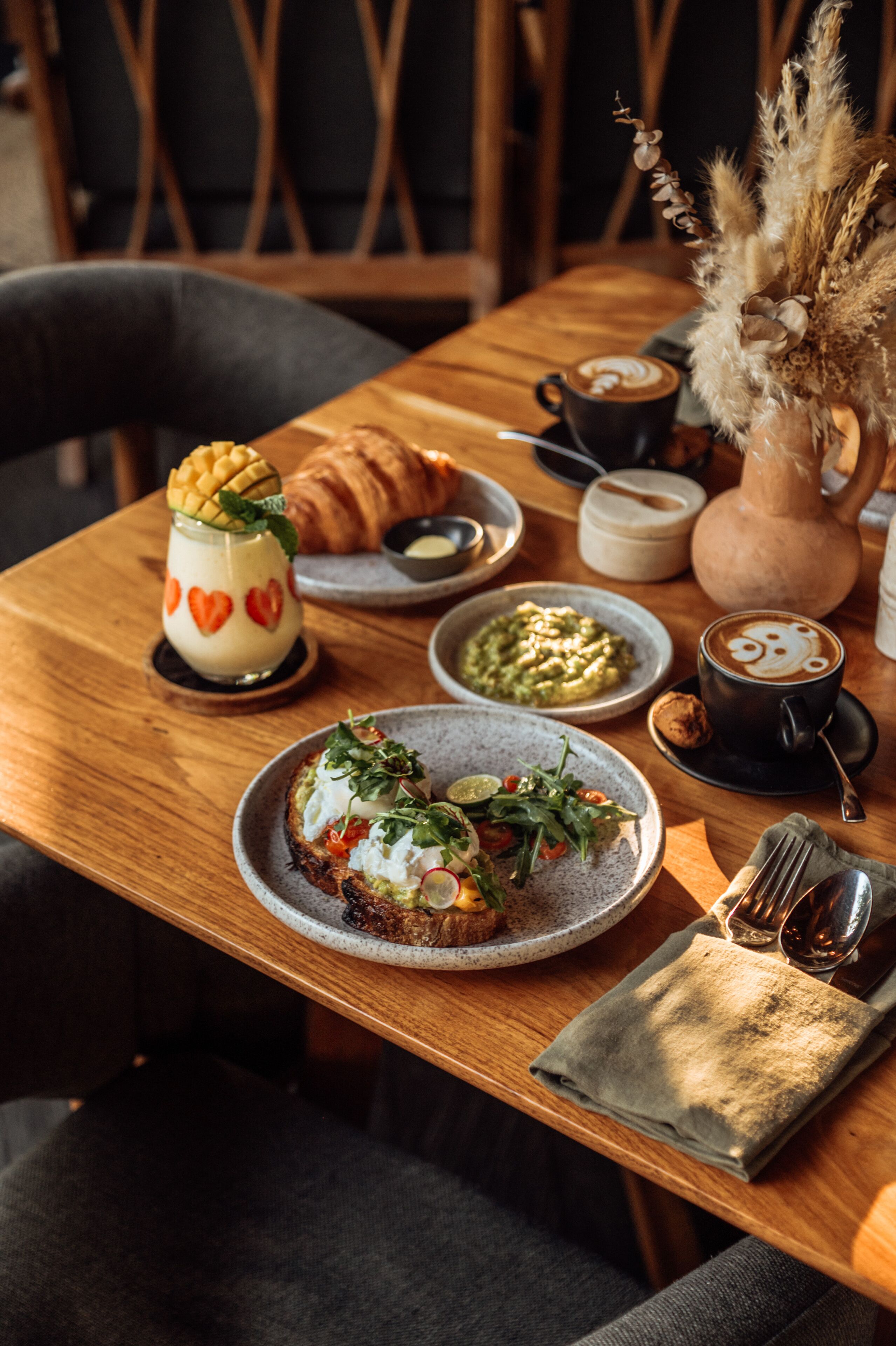 Une table de salle à manger en bois, avec une assiette de nourriture, une tasse de café et un vase contenant des plantes séchées.