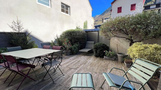 A wooden deck with a table and chairs, surrounded by potted plants and a building with windows.