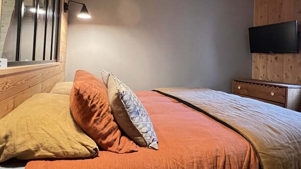 Une chambre à coucher avec un lit, des oreillers, une lampe de chevet, une commode et un téléviseur fixé au mur.