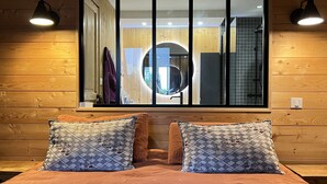 A bedroom with a large window, wooden walls, a bed with patterned pillows, and two wall-mounted lamps.