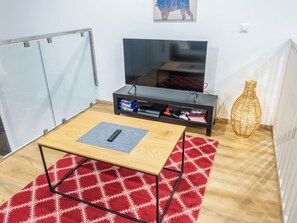 Una habitación con televisión, una pequeña mesa con control remoto, una estantería con juegos y un jarrón decorativo.