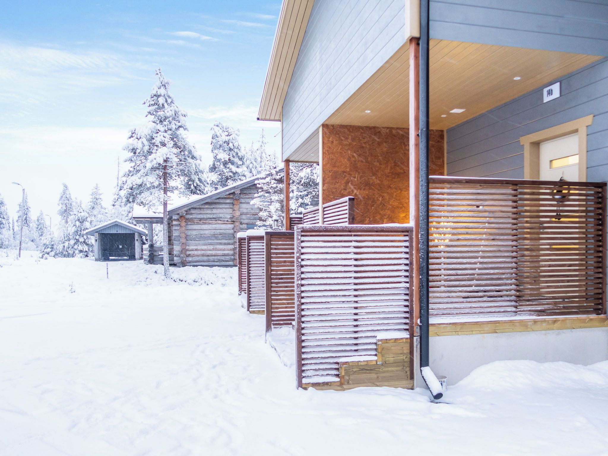 A snow-covered outdoor area with a wooden fence, a building with a wooden facade, and snow-covered trees.