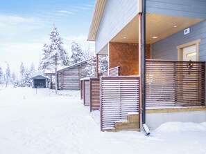 A snow-covered outdoor area with a wooden fence, a building with a wooden facade, and snow-covered trees.
