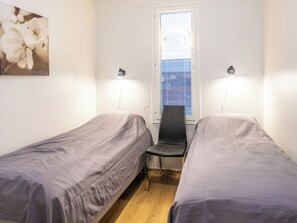 Quarto com duas camas de solteiro, uma cadeira, janela com persianas e luminária na parede.