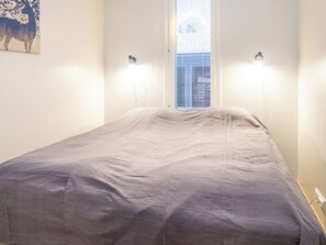 Una cama individual con cubrecolcha gris en una habitación con paredes blancas y una ventana con persianas.