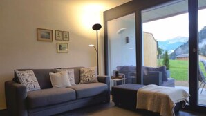 A modern living room with a grey sofa, two side tables, and a large sliding glass door leading to an outdoor area with a view of mountains. - Greenview by Interhome (Blankenburg)