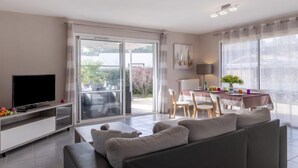Ein modernes Wohnzimmer mit Fernseher, einer Couch, einer Essecke mit Tisch und Stühlen und großen Fenstern mit Blick ins Freie.