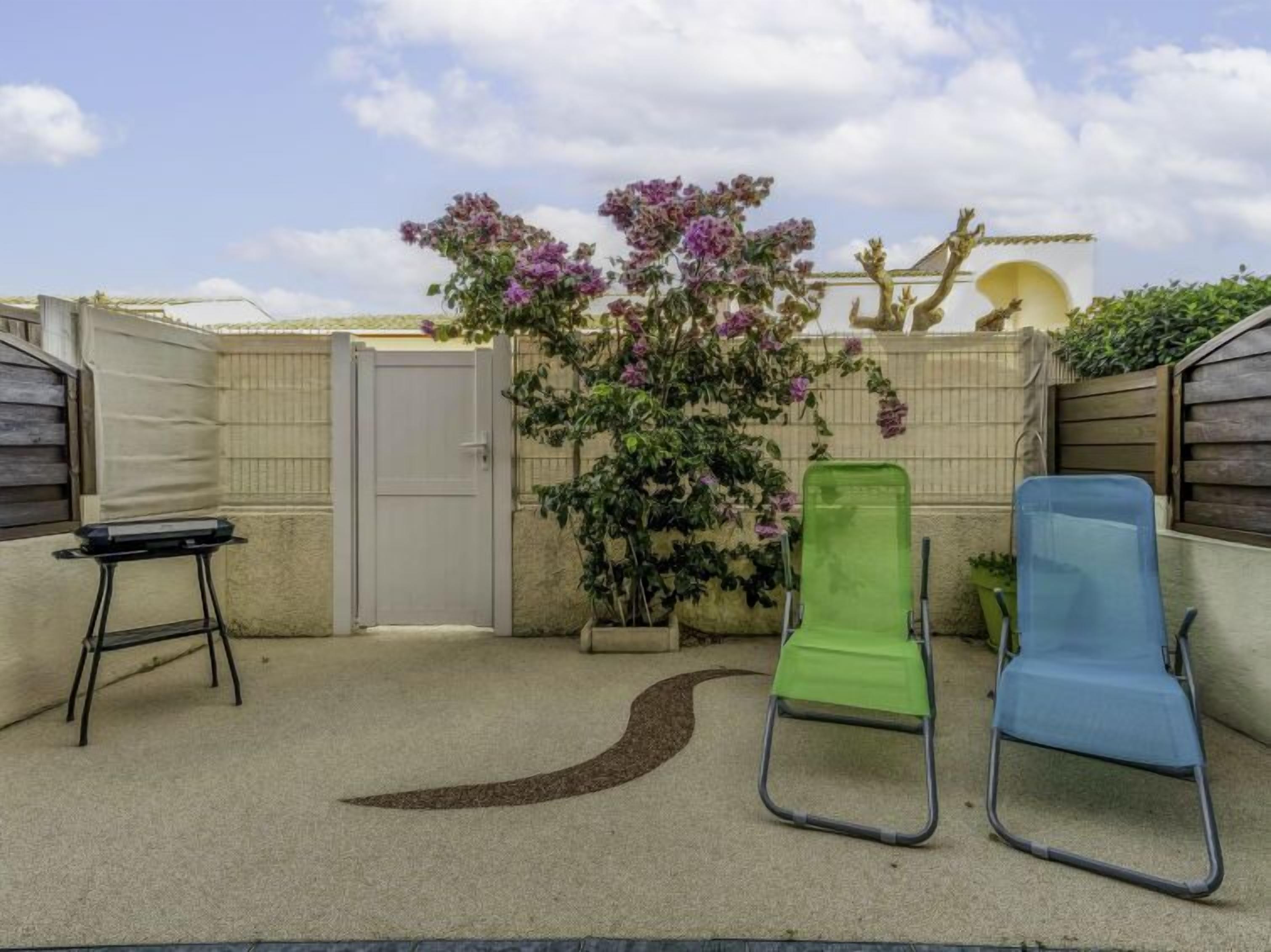 Un petit espace extérieur avec une porte blanche, une chaise verte et une chaise bleue, ainsi qu’une plante à fleurs violettes.