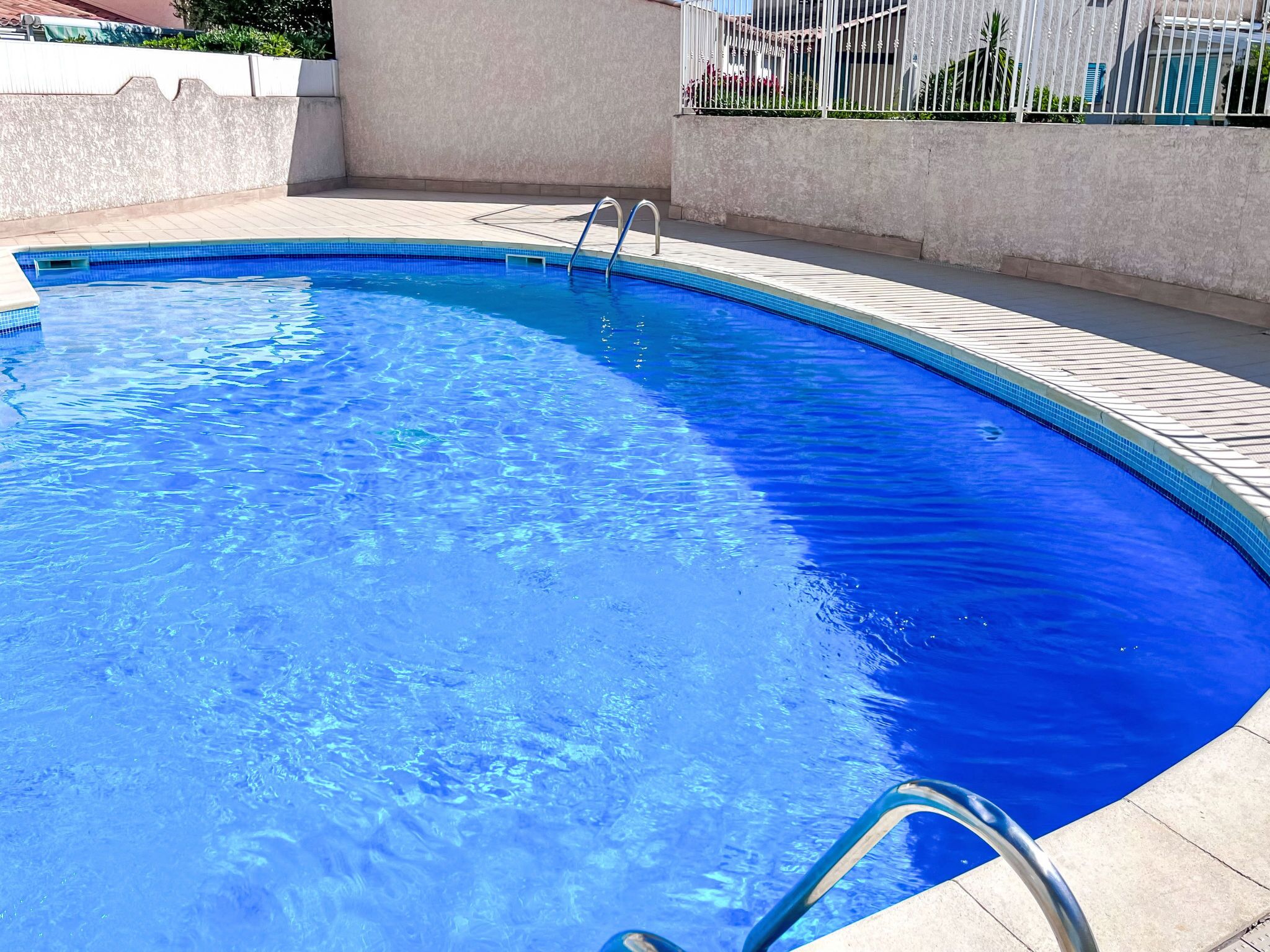 A clear blue swimming pool with a metal ladder leading into it, surrounded by a tiled deck and a low white wall.